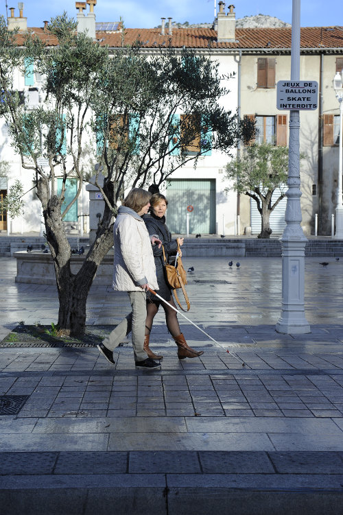 Deux femmes marchent dans une rue ensoleillée. L'une s'aide d'une canne blanche pour se déplacer. (Agrandir l'image)